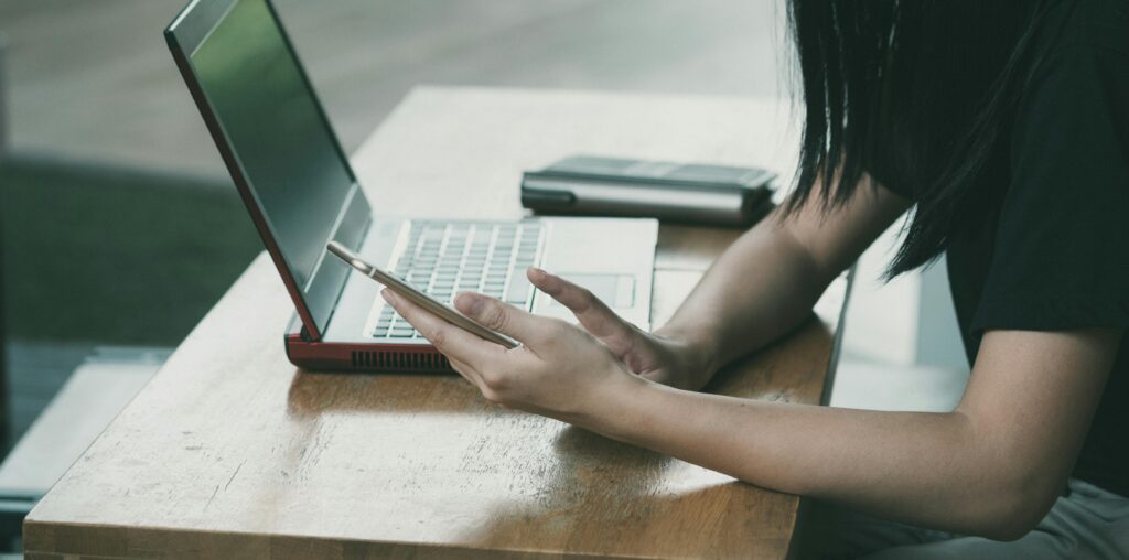 A young woman online part time jobs with her smartphone and laptop at a table indoors.