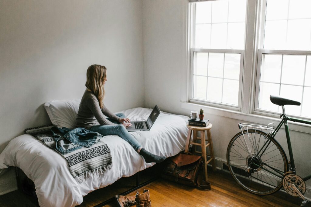 A young woman works on a laptop in a cozy, minimalist bedroom setting, showcasing remote work lifestyle and finding difference between affiliate marketing and digital